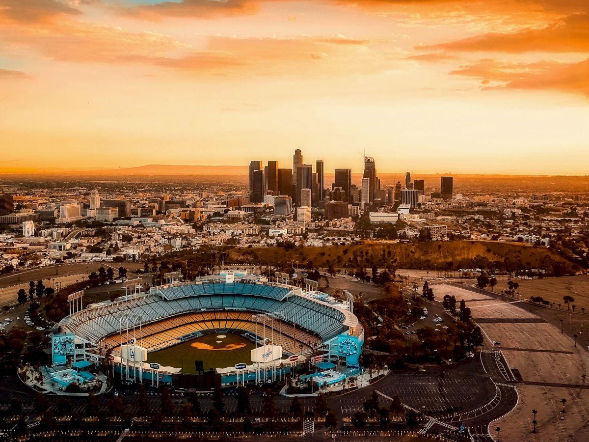 An aerial view of a baseball stadium with a city skyline in the background during sunset. The sky is a mix of orange and yellow hues.