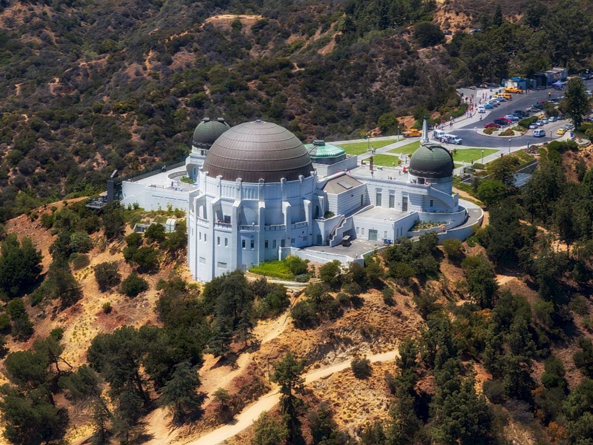 An aerial view of a large observatory with a central dome and two smaller domes, situated amidst hilly terrain surrounded by trees and a parking area.